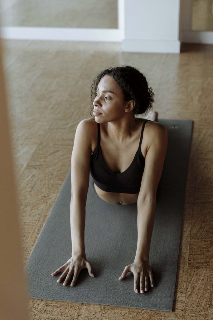 A woman in a black sports bra doing a yoga stretch on a gray mat indoors on a wooden floor