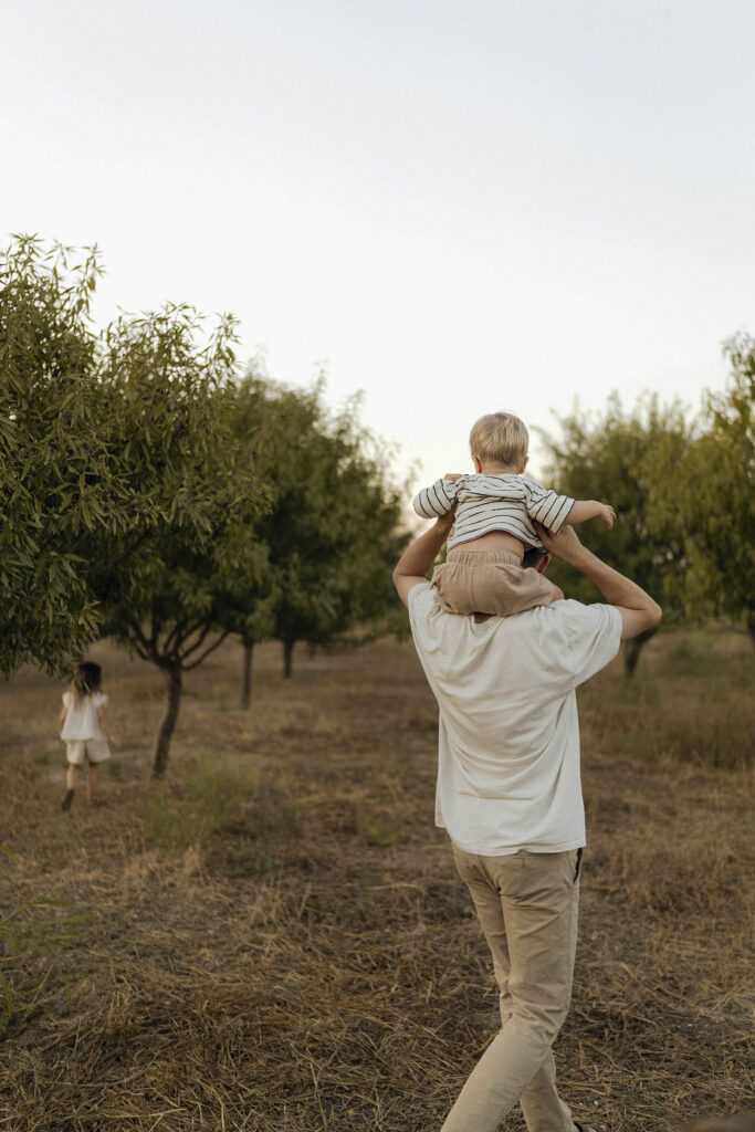 An adult carries a child on their shoulders while walking through an orchard_ another child walks in the background among the trees
